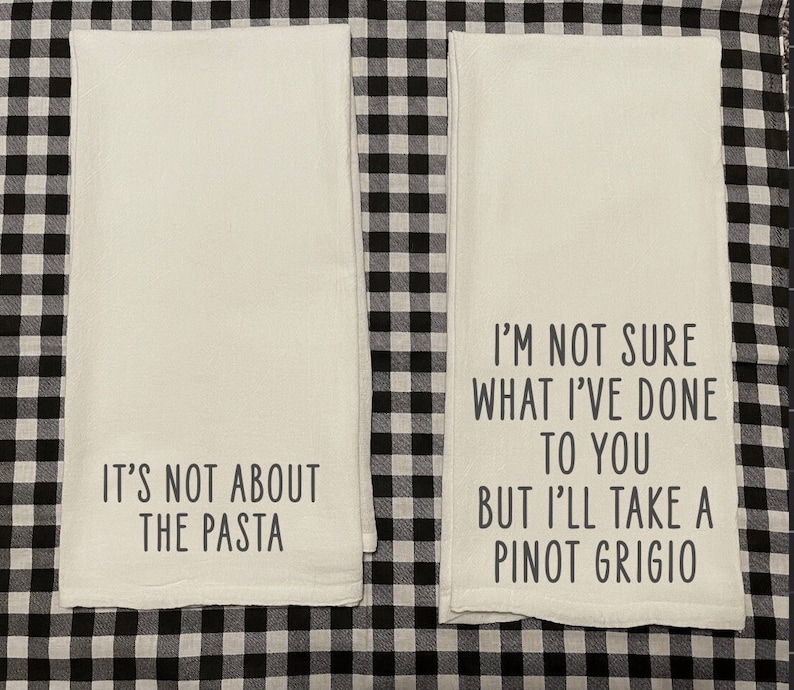May include: Two white kitchen towels with black text. The first towel says "It's not about the pasta". The second towel says "I'm not sure what I've done to you but I'll take a Pinot Grigio".