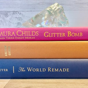 May include: A stack of three hardcover books. The top book is pink with gold lettering that reads "LAURA CHILDS GLITTER BOMB with TERRIE FARLEY MORAN". The middle book is orange with gold lettering. The bottom book is blue with gold lettering that reads "G. J. MEYER | The WORLD REMADE".