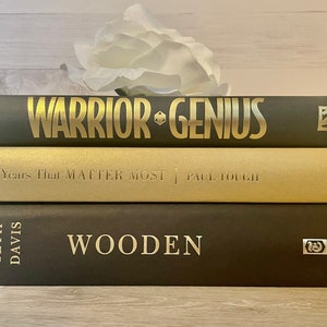 May include: A stack of three hardcover books with gold lettering on black covers. The top book is titled "Warrior Genius" by Dimartino. The middle book is titled "The Years That Matter Most" by Paul Tough. The bottom book is titled "Wooden" by Seth Davis.