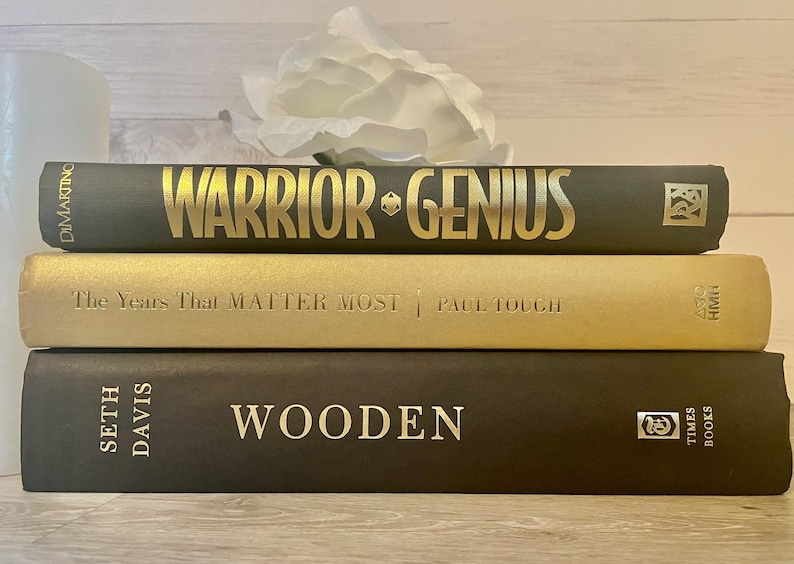 May include: A stack of three hardcover books with gold lettering on black covers. The top book is titled "Warrior Genius" by Dimartino. The middle book is titled "The Years That Matter Most" by Paul Tough. The bottom book is titled "Wooden" by Seth Davis.