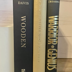 May include: Three hardcover books with black covers and gold lettering. The book on the left is titled "Wooden" by Seth Davis. The middle book is titled "The Years That Matter Most" by Paul Tough. The book on the right is titled "Warrior Genius" by Dimartino.