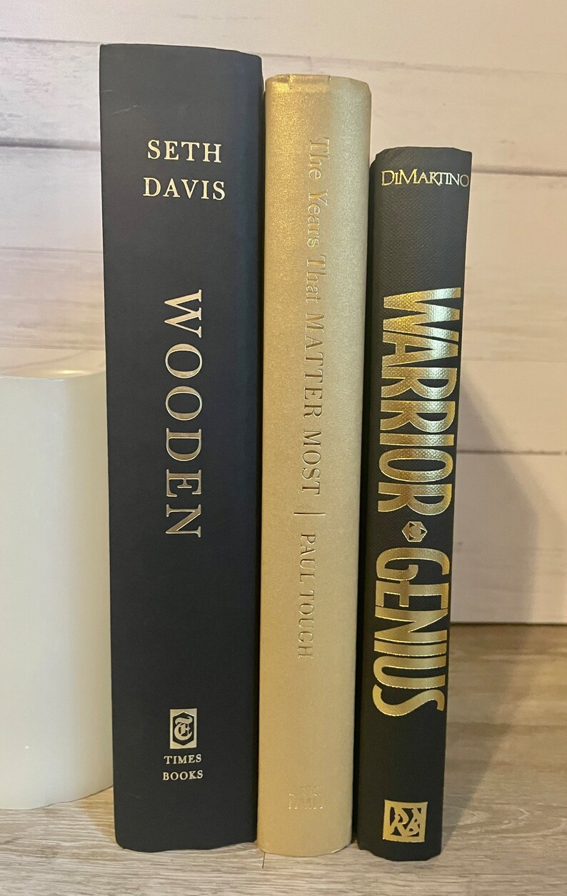 May include: Three hardcover books with black covers and gold lettering. The book on the left is titled "Wooden" by Seth Davis. The middle book is titled "The Years That Matter Most" by Paul Tough. The book on the right is titled "Warrior Genius" by Dimartino.