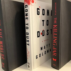 May include: Three hardback books on a white shelf. The middle book is white with red splatter and the title "Gone to Dust" by Matt Goldman. The book on the left is black with the title "Defy the Worlds". The book on the right is black with the title "The Killing Jar" by Bosworth.