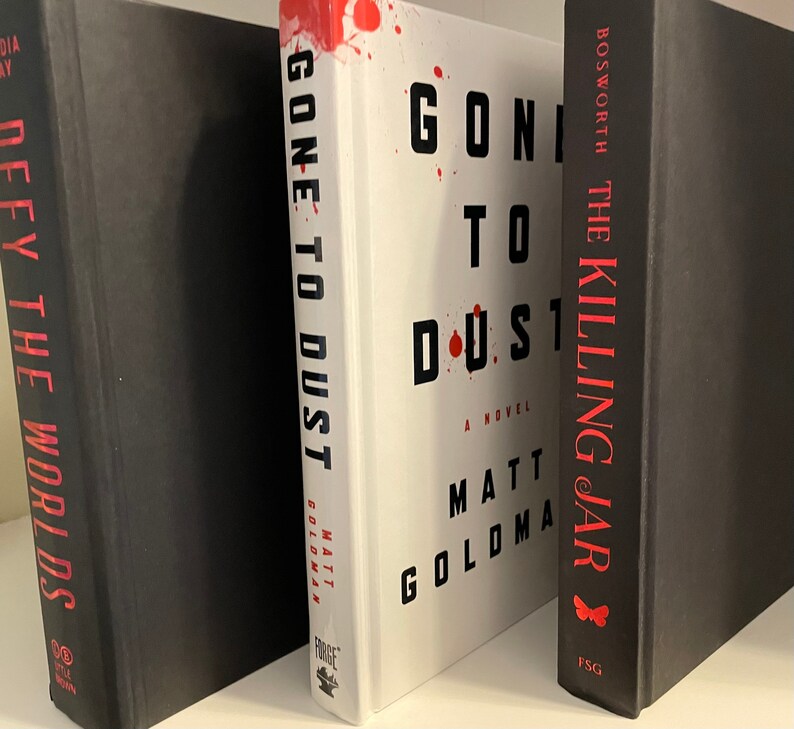 May include: Three hardback books on a white shelf. The middle book is white with red splatter and the title "Gone to Dust" by Matt Goldman. The book on the left is black with the title "Defy the Worlds". The book on the right is black with the title "The Killing Jar" by Bosworth.