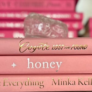 May include: A stack of pink hardcover books with gold and silver lettering. The titles include "Coyote Lost and Found", "honey", and "Tell Me Everything". A clear, textured object sits atop the stack, with more pink books in the background.
