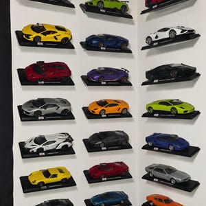 May include: A collection of twenty-one miniature Lamborghini model cars displayed on individual black rectangular stands against a white wall. The cars feature various colours including red, yellow, blue, green, white, silver, orange, and black.
