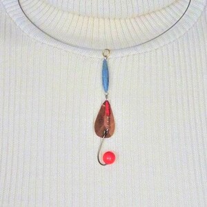 Puede incluir: Un collar de cadena plateada con un colgante de señuelo de pesca de cobre. El señuelo es rojo, azul y marrón con una cuenta roja en la parte inferior.