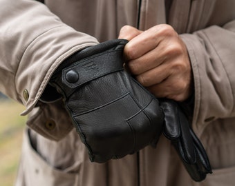 May include: A pair of black leather gloves with a button fastening on the wrist. The gloves have a textured surface and are being held by a person.