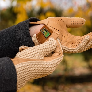 May include: A pair of tan leather gloves with a knitted cuff. The gloves have a perforated design and a small leather patch with a logo on the wrist.