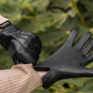 Op de afbeelding: Een paar zwarte leren handschoenen worden aangetrokken. De handschoenen hebben een klassiek ontwerp met stiksels.