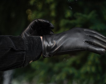 May include: A pair of black leather gloves with a ribbed cuff. The gloves are being worn by a person with a black corduroy jacket.