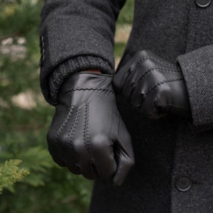 Può includere: Un paio di guanti in pelle nera con dettagli di cucitura. I guanti sono indossati da una persona che indossa un cappotto scuro.