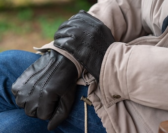 May include: A pair of black leather gloves with stitching detail. The gloves are folded and resting on a person's lap.