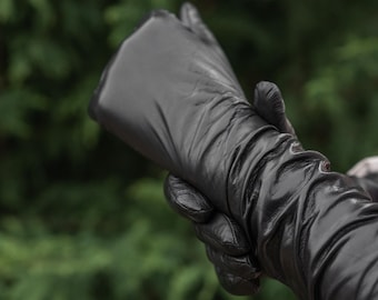 May include: Close-up of a black leather glove, with a long, gathered sleeve. The glove is made of a shiny, smooth leather material. The background is a blurred green, suggesting an outdoor setting.