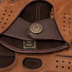 Puede incluir: Primer plano de guantes de conducir de cuero marr&oacute;n con un dise&ntilde;o perforado. Los guantes tienen una correa de cuero marr&oacute;n con un bot&oacute;n de lat&oacute;n que dice "Gloves". La etiqueta de los guantes dice "PECS".