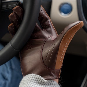 Puede incluir: Un guante de conducir de cuero marr&oacute;n con una palma de ante beige y detalles perforados. El guante se lleva en la mano de una persona, sujetando un volante.