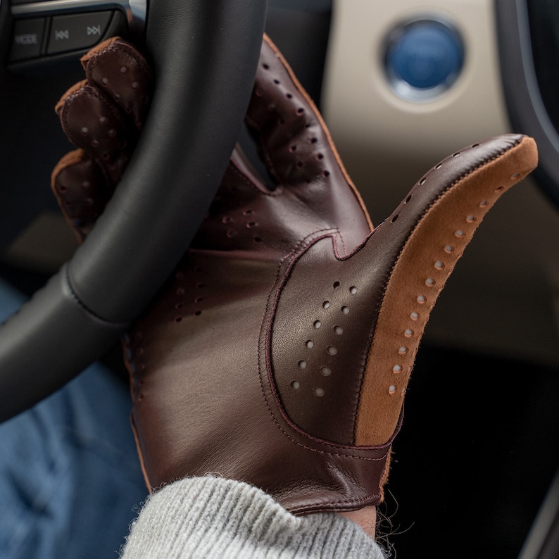 Puede incluir: Un guante de conducir de cuero marr&oacute;n con una palma de ante beige y detalles perforados. El guante se lleva en la mano de una persona, sujetando un volante.