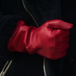 Könnte beinhalten: Ein Paar rote Lederhandschuhe mit einem strukturierten Design. Die Handschuhe sind gefaltet und werden in einer Hand gehalten.