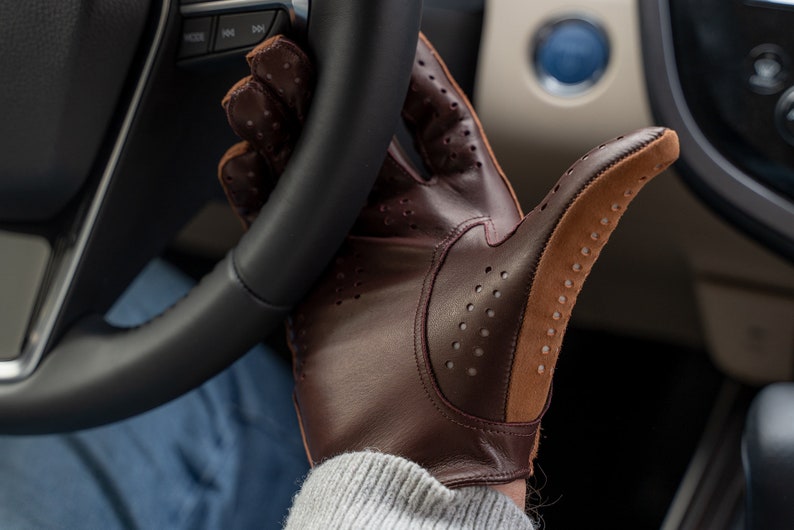 Puede incluir: Un par de guantes de conducir de cuero marr&oacute;n con detalles perforados y una palma de gamuza. Los guantes se llevan en la mano de una persona, agarrando un volante.