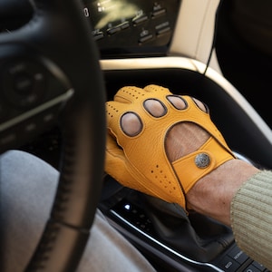 Può includere: Primo piano di una mano che indossa un guanto da guida in pelle gialla con perforazioni e un dettaglio a bottone. Il guanto è sul cambio di un'auto.