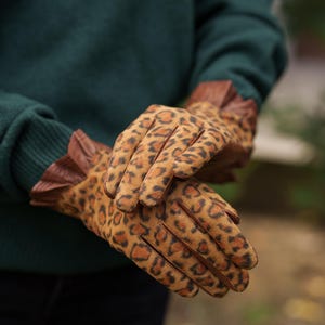 Könnte beinhalten: Ein Paar Handschuhe mit Leopardenmuster. Die Handschuhe sind beige mit braunen Flecken und haben braune Lederbesätze. Getragen von einer Person, die einen dunkelgrünen Pullover trägt.