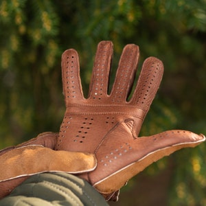Pu&ograve; includere: Guanti da guida in pelle marrone con un design traforato. I guanti hanno un polsino in pelle marrone e sono mostrati con le dita estese.