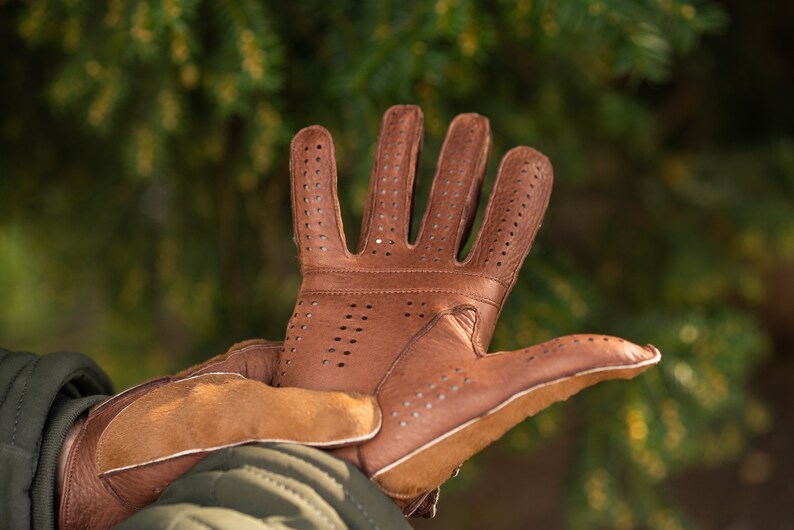 Pu&ograve; includere: Guanti da guida in pelle marrone con un design traforato. I guanti hanno un polsino in pelle marrone e sono mostrati con le dita estese.