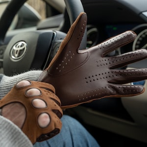 Puede incluir: Un par de guantes de conducir de cuero marr&oacute;n con detalles perforados. Los guantes los lleva una persona en un coche.