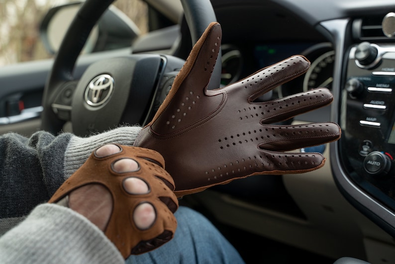 Puede incluir: Un par de guantes de conducir de cuero marr&oacute;n con detalles perforados. Los guantes los lleva una persona en un coche.