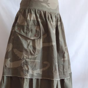 May include: A khaki camouflage print skirt. The skirt has a high waist, a large pocket, and a ruffled hem. The fabric is a blend of green, brown, and tan. It has a zip closure. The skirt is a midi length, suitable for various occasions.