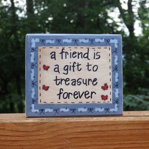May include: A blue wooden sign with a white border and red hearts. The sign reads "A friend is a gift to treasure forever".