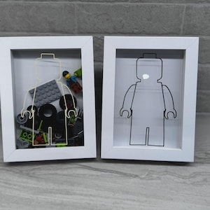 May include: Two white shadow box frames. One frame contains a yellow outline of a Lego figure filled with Lego bricks and a Lego figure. The other frame has a black outline of a Lego figure.