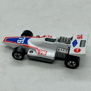 May include: A white and red vintage toy race car with the number "76" in blue and red. The car has black wheels and a silver engine detail. The word "BELL" is visible on the rear wing.