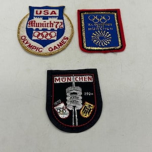May include: Three embroidered patches from the 1972 Munich Olympic Games. One shield-shaped patch reads USA Munich 72 Olympic Games. A rectangular blue patch reads XX Olympiade 1972 Munchen. A black patch features the Munich TV tower with 290m text.