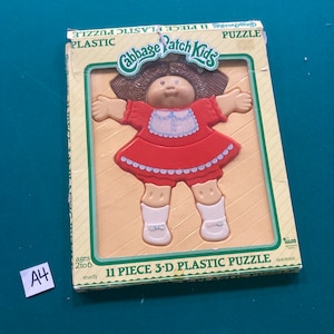 May include: An 11-piece 3-D plastic puzzle of a Cabbage Patch Kid doll. The doll is wearing a red dress with white trim and white shoes. The puzzle is for ages 2 to 6 and is sturdy and non-toxic.