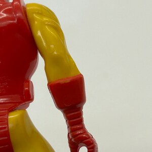 May include: Close-up of a red and yellow plastic action figure's right arm. The arm is bent at the elbow and the hand is closed into a fist. The figure's arm is detailed with red and yellow paint.