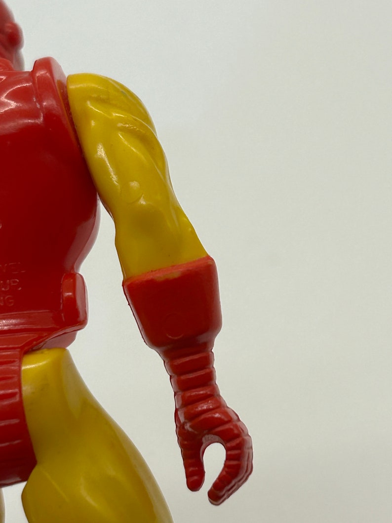 May include: Close-up of a red and yellow plastic action figure's right arm. The arm is bent at the elbow and the hand is closed into a fist. The figure's arm is detailed with red and yellow paint.