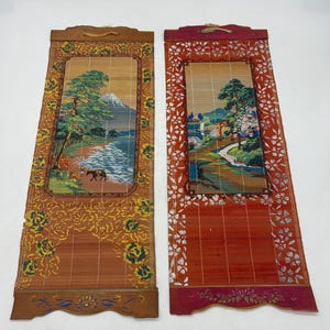 May include: Two decorative bamboo wall hangings with Japanese landscape scenes. One features a mountain and river, the other a path and traditional gate. Both have ornate borders in shades of orange and red, with wooden tops.