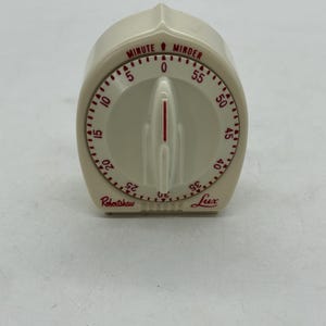 May include: A vintage off-white kitchen timer with red numerals and markings. The timer, labeled "Minute Minder" and "Lux" by Robertshaw, features a central dial and a needle for setting the time. The timer's face displays numbers from 0 to 55.