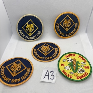 May include: Five embroidered patches with various designs and text. The patches are circular and feature a blue, yellow, and green color scheme. The text on the patches includes "Pack Committee", "Assistant Cubmaster", "Assistant Den Leader", "Super Pop Sales Award", and "1989".