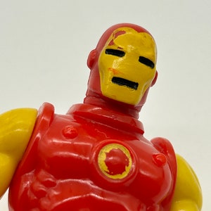 May include: A red and yellow plastic action figure of Iron Man. The figure has a red torso and arms, a yellow head and shoulders, and a yellow circle on the chest. The figure's face is painted yellow with black eyes and a black mouth.