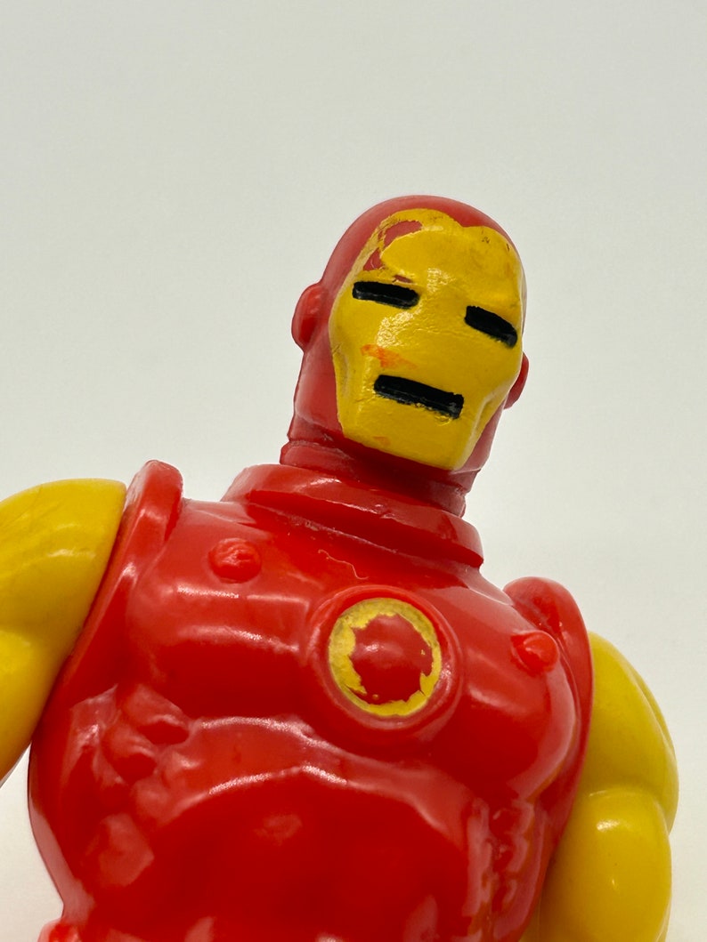 May include: A red and yellow plastic action figure of Iron Man. The figure has a red torso and arms, a yellow head and shoulders, and a yellow circle on the chest. The figure's face is painted yellow with black eyes and a black mouth.
