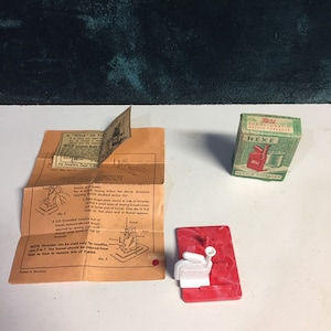 May include: A red and white plastic needle threader with a small white handle and a red base. The threader is labeled "HEXE" and is packaged in a small box with the text "The Automatic Needle Threader". The box is also labeled "HEXE". The image also includes instructions for using the threader.