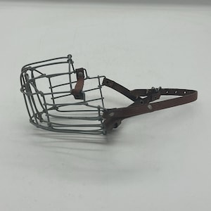 May include: A wire dog muzzle with a cage-like front and brown leather straps. The muzzle is made of gray metal wires and has a brown leather strap that goes around the dog's head. The muzzle is designed to prevent biting.