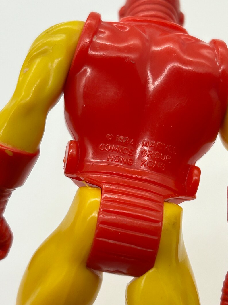 May include: A red and yellow plastic action figure of Iron Man. The figure is standing with its back to the camera. The figure has a copyright notice on the back that reads "&copy; 1984 MARVEL COMICS GROUP HONG KONG."