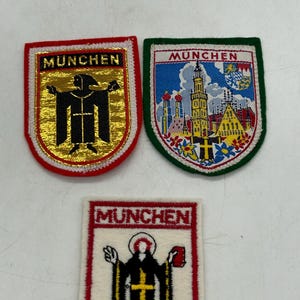 May include: Three embroidered patches featuring the word MUNCHEN. One patch has a gold background with a black monk figure and red border. Another shows a colorful city scene with a green border. The third is white with a black monk figure.