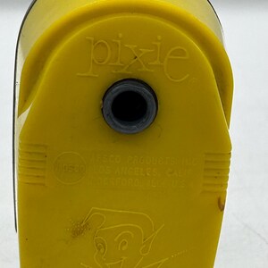 May include: A bright yellow manual pencil sharpener with a rounded top. The front features the embossed text "pixie" above a circular opening, and "Apsco Products Inc. Los Angeles, Calif. Rockford, Ill. U.S.A." with a small jester graphic below.