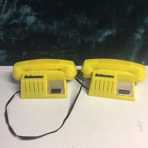 May include: Two yellow toy telephones with black cords. The phones have a black button and a speaker on the front. The text "Telephone" is printed on the front of each phone.