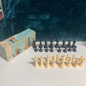 May include: A vintage chess set with black and white plastic pieces. The pieces are arranged on a white surface. The box for the chess set is in the upper left corner of the image. The box is blue and white and says "Chess Men" on the front.
