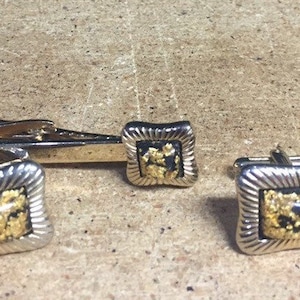 May include: A set of gold and silver cufflinks and a tie clip. The cufflinks and tie clip feature a black background with gold flecks.